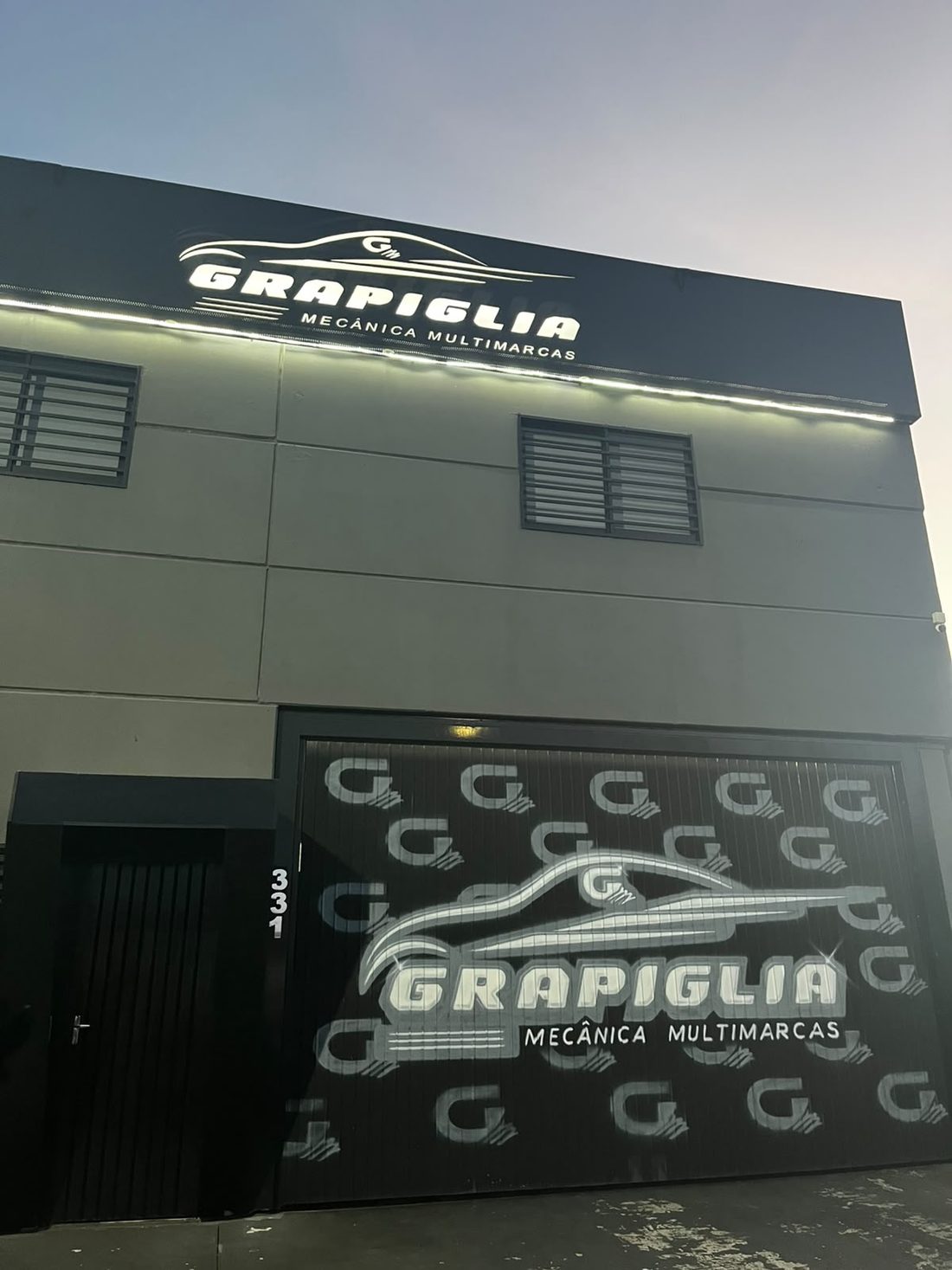 Entrada da oficina ao anoitecer, portão com a arte da marca